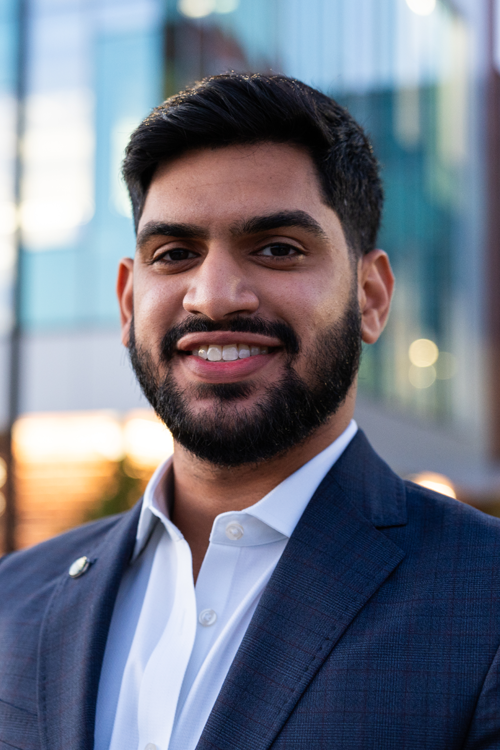 Man wearing a blue suit jacked and blue shirt looking at camera and smiling. He is standing outside in front of buildings.